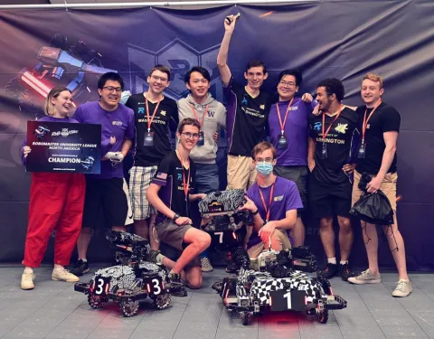 Ten students, several wearing purple, stand in a group with two small wheeled robots in front of them