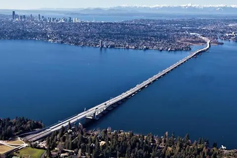 Aerial view, looking west, of the new SR 520 floating bridge, with the Medina shoreline in the foreground and downtown Seattle skyline in the distance.