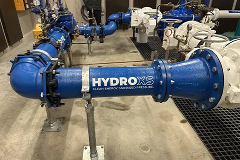 Blue industrial pipe labeled "HYDRO XS" in a facility with various pipes and machinery