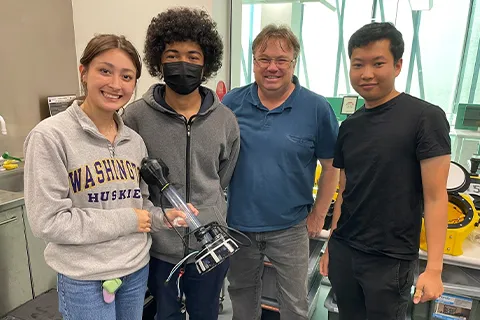 Three students and one faculty member stand in a lab setting. One student holds an arm attachment for a robot.