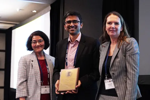 Aniruddh Vashisth is smiling and holding a plaque, standing between two people at the ASC conference.