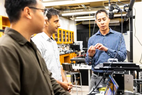 Three researchers in the Molecular Biophotonics Lab stand around a table. On the table are 3D microscopy technologies.