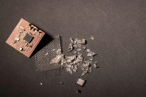 A small brown circuit board. To its right are a small copper plate, sheets of glass fibers, small chunks of vitrimer plastic and a computer chip. 