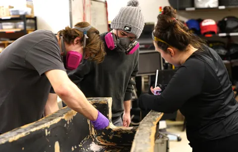 Two students wearing large masks are sanding and examining a large structure that will be part of the Formula car. A third student is taking notes.