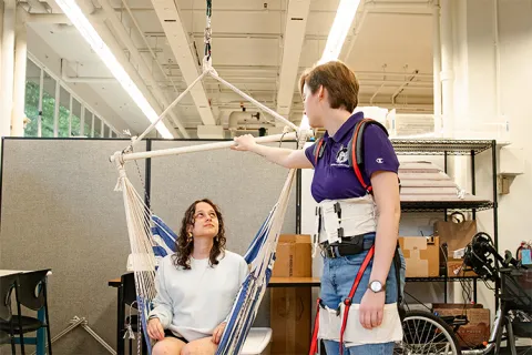 One person is sitting in a hammock chair, while another person holds part of the frame structure.