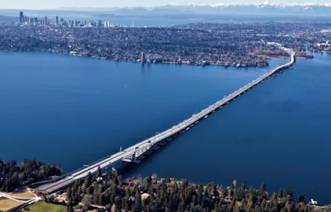 Aerial view, looking west, of the new SR 520 floating bridge, with the Medina shoreline in the foreground and downtown Seattle skyline in the distance.