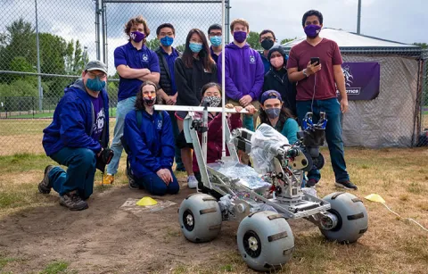 A crowd of 12 people in masks stand in a group outdoors behind a robot that looks like a mars rover