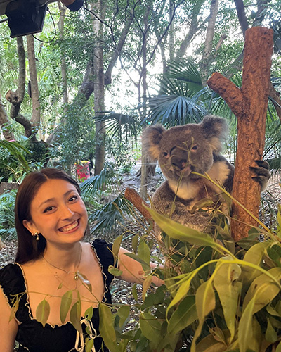 Madeline Beltran smiles while standing next to a koala in a tree.