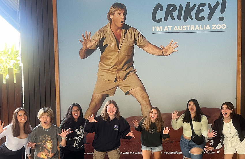 A group of students with their hands up stand underneath a sign that reads Crikey, I'm at the Australia Zoo.