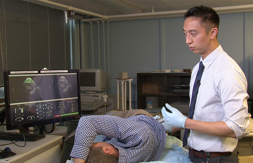 A man lies down in a hospital bed while another man holds a handheld device to his back while seeing ultrasound imaging on a computer screen.