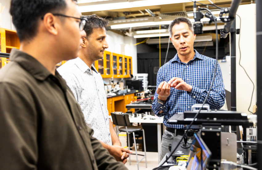 Three researchers in the Molecular Biophotonics Lab stand around a table. On the table are 3D microscopy technologies.