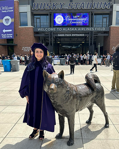 Em Triolo wearing a graduation robe and standing next to a statue of a husky dog outside of the Husky Stadium.