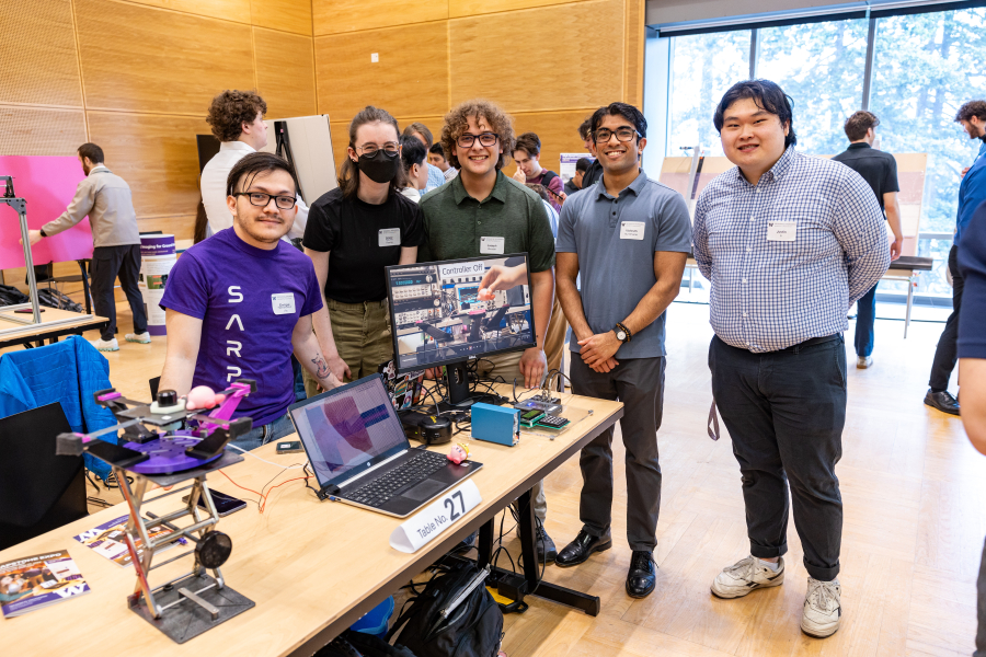 Five students stand near their table that showcases their platform that stabilizes vibrations and screens showing a video demo.