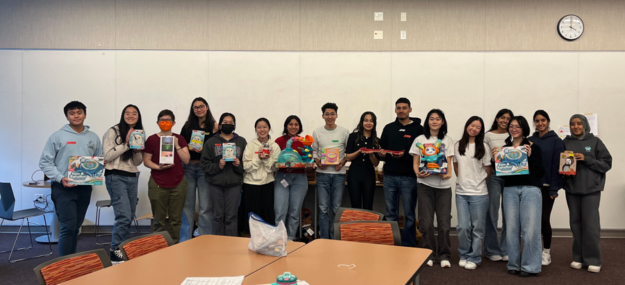 Sixteen students at the HuskyADAPT toy adaptation event stand in front of a whiteboard holding various toys.