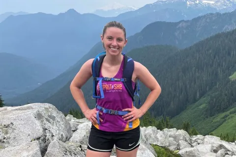 A young woman stands atop a mountain in athletic gear with a backdrop of mountains