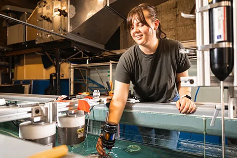 Woman standing next to a metal railing, placing an object inside a flume at the Harris Hydraulics Lab