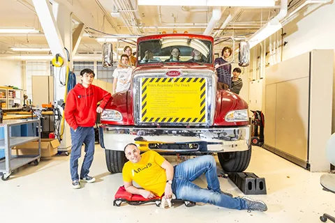 A group of people posing to the camera on a red semi-truck in a workshop. 