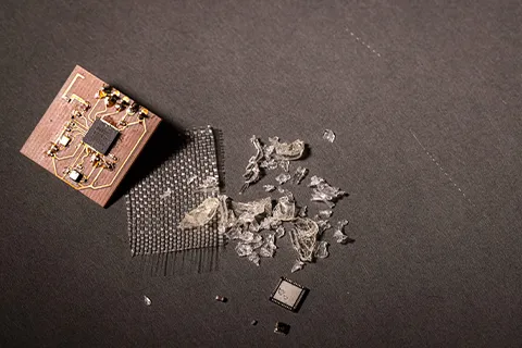 A small brown circuit board sits on a gray background. To its right are a small copper plate, sheets of glass fibers in a crosshatch pattern, small chunks of vitrimer plastic that’s been removed from a circuit board, and a computer chip. 