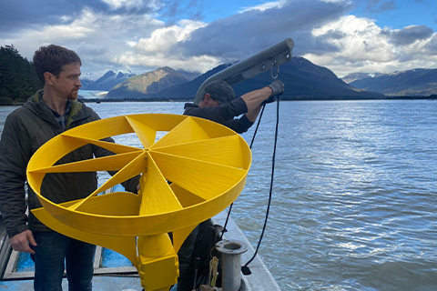 Two men on a boat. One man holds a large yellow fan-shaped object while the other adjusts a rope connected to a boom arm.