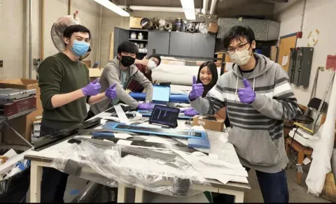 Five masked and gloved individuals giving thumbs-up around a table with devices in a workshop.