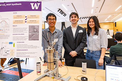Three students stand behind a vessel that is a reactor for synthesizing materials.