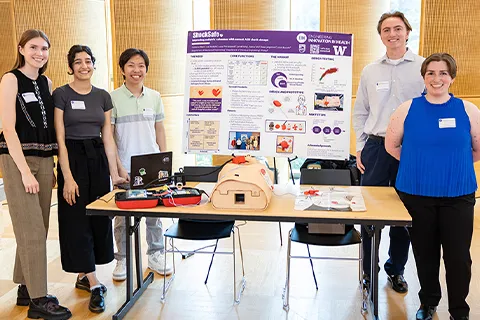 Five ShockSafe team members stand next to their poster and their prototype of their device.